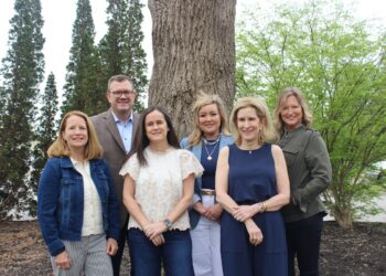 A group of six professionally dressed adults—five women and one man—pose together outdoors in front of a thick, textured tree trunk. They stand close together smiling, with tall evergreen trees and leafy green bushes creating a natural background.