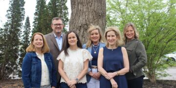 A group of six professionally dressed adults—five women and one man—pose together outdoors in front of a thick, textured tree trunk. They stand close together smiling, with tall evergreen trees and leafy green bushes creating a natural background.