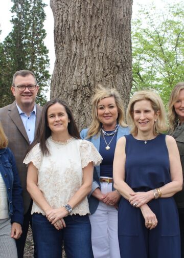 A group of six professionally dressed adults—five women and one man—pose together outdoors in front of a thick, textured tree trunk. They stand close together smiling, with tall evergreen trees and leafy green bushes creating a natural background.