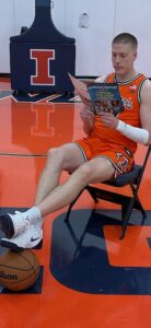 An eye-level shot of an Illinois Fighting Illini men's basketball player sitting on a black folding chair in the center of an orange basketball court. He is wearing a bright orange uniform with black and white trim, and his left forearm is wrapped in white athletic tape. He holds open a copy of a magazine titled "Kokomo-Tipton," focusing on an article inside. His white Nike sneakers rest directly on top of a leather basketball on the polished floor. In the background, a blue padding structure features a prominent orange and white "I" logo.