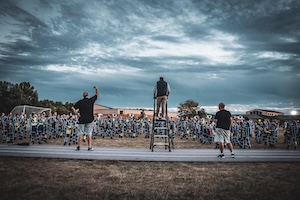 A wide-angle, low-angle shot from behind three directors overseeing a large marching band practice on a field. One director stands atop a tall ladder in the center, while the other two stand on a paved track in the foreground, one with an arm raised. The massive band, dressed in blue and white uniforms, is spread across the grass field in the midground. The scene is set under a dramatic, cloudy twilight sky with cool blue and grey tones.
