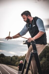 A side-profile shot shows a man with a dark beard standing atop a black ladder on a track. He is dressed in a black and grey hooded shirt and tan pants, focused on the task at hand. He holds a pair of drumsticks, one in each hand, and appears to be keeping time or conducting. In the background, another person is visible at the bottom of the ladder, and a paved track stretches into the distance under a pale, overcast sky.