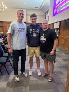 An indoor full-body portrait shows three men standing together and smiling in a large meeting hall or banquet room. The man on the left wears a light blue t-shirt with "MOPO" in large blue letters and dark pants. The young man in the center wears a navy blue "FCA" (Fellowship of Christian Athletes) hoodie and tan shorts. The young man on the right wears a black t-shirt that says "WE REIGN SUPREME" and grey athletic shorts. They are standing on a grey patterned carpet in front of a wood-paneled wall. To the right, a large purple presentation screen is visible, and to the left, people sit at tables under a chandelier.