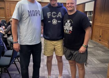 An indoor full-body portrait shows three men standing together and smiling in a large meeting hall or banquet room. The man on the left wears a light blue t-shirt with "MOPO" in large blue letters and dark pants. The young man in the center wears a navy blue "FCA" (Fellowship of Christian Athletes) hoodie and tan shorts. The young man on the right wears a black t-shirt that says "WE REIGN SUPREME" and grey athletic shorts. They are standing on a grey patterned carpet in front of a wood-paneled wall. To the right, a large purple presentation screen is visible, and to the left, people sit at tables under a chandelier.