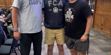 An indoor full-body portrait shows three men standing together and smiling in a large meeting hall or banquet room. The man on the left wears a light blue t-shirt with "MOPO" in large blue letters and dark pants. The young man in the center wears a navy blue "FCA" (Fellowship of Christian Athletes) hoodie and tan shorts. The young man on the right wears a black t-shirt that says "WE REIGN SUPREME" and grey athletic shorts. They are standing on a grey patterned carpet in front of a wood-paneled wall. To the right, a large purple presentation screen is visible, and to the left, people sit at tables under a chandelier.