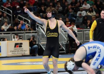 A high school wrestler wearing a black and gold singlet with "Cowan" printed across the front smiles and points one finger upward in celebration on a blue and yellow wrestling mat. He wears black headgear around his neck, while an opposing wrestler in a blue singlet labeled "Trojans" sits on the mat in the blurred foreground. A coach in a black vest looks on from the background next to an Indiana Farm Bureau Insurance sign.