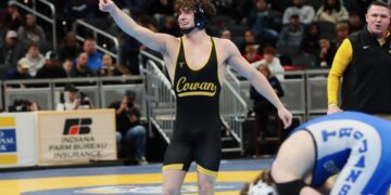 A high school wrestler wearing a black and gold singlet with "Cowan" printed across the front smiles and points one finger upward in celebration on a blue and yellow wrestling mat. He wears black headgear around his neck, while an opposing wrestler in a blue singlet labeled "Trojans" sits on the mat in the blurred foreground. A coach in a black vest looks on from the background next to an Indiana Farm Bureau Insurance sign.