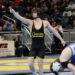 A high school wrestler wearing a black and gold singlet with "Cowan" printed across the front smiles and points one finger upward in celebration on a blue and yellow wrestling mat. He wears black headgear around his neck, while an opposing wrestler in a blue singlet labeled "Trojans" sits on the mat in the blurred foreground. A coach in a black vest looks on from the background next to an Indiana Farm Bureau Insurance sign.