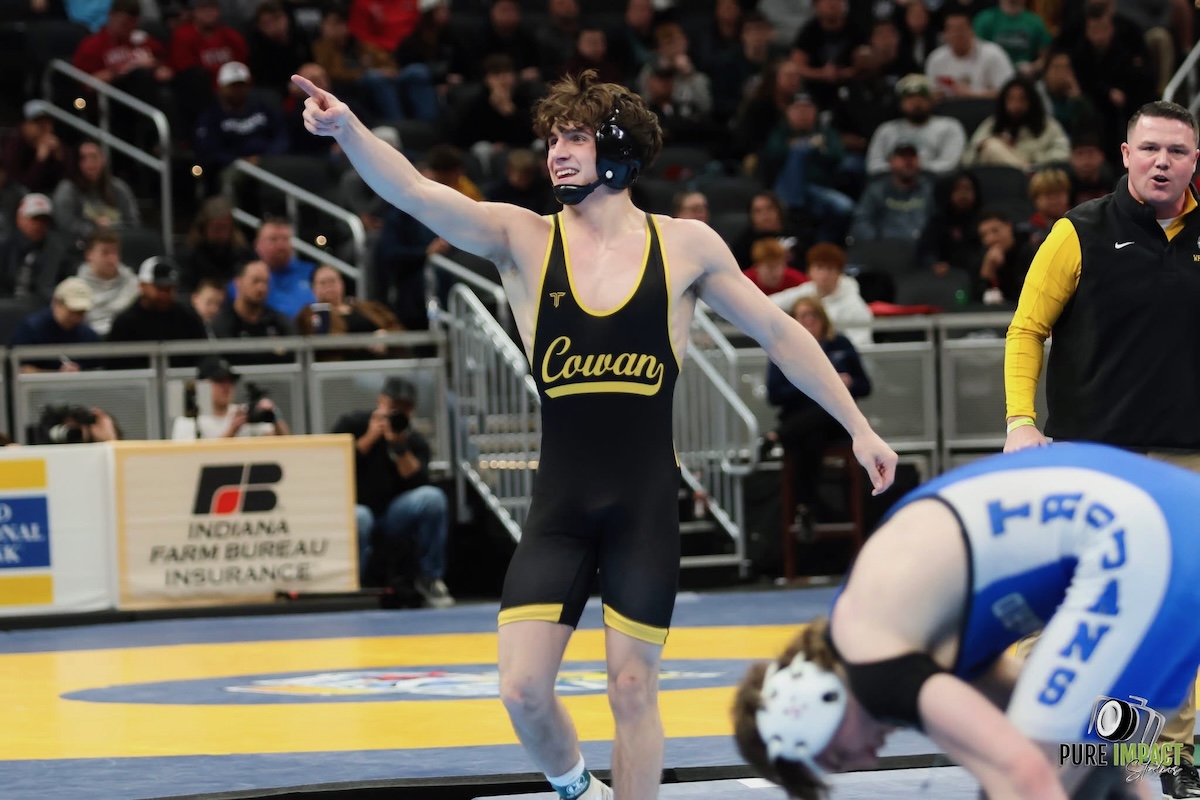 A high school wrestler wearing a black and gold singlet with "Cowan" printed across the front smiles and points one finger upward in celebration on a blue and yellow wrestling mat. He wears black headgear around his neck, while an opposing wrestler in a blue singlet labeled "Trojans" sits on the mat in the blurred foreground. A coach in a black vest looks on from the background next to an Indiana Farm Bureau Insurance sign.