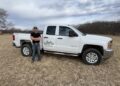 A man stands with his arms crossed next to a white Chevrolet Silverado 2500 HD pickup truck parked in a dry, grassy field. The man wears a black t-shirt, blue jeans, and a baseball cap, smiling at the camera. The front door of the truck features a custom logo that reads "Jacks Lawn Service" along with a phone number. In the background, a line of bare trees sits under an overcast sky with textured white and gray clouds.