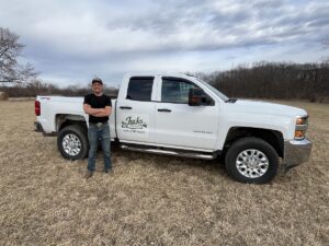 A man stands with his arms crossed next to a white Chevrolet Silverado 2500 HD pickup truck parked in a dry, grassy field. The man wears a black t-shirt, blue jeans, and a baseball cap, smiling at the camera. The front door of the truck features a custom logo that reads "Jacks Lawn Service" along with a phone number. In the background, a line of bare trees sits under an overcast sky with textured white and gray clouds.