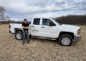 A man stands with his arms crossed next to a white Chevrolet Silverado 2500 HD pickup truck parked in a dry, grassy field. The man wears a black t-shirt, blue jeans, and a baseball cap, smiling at the camera. The front door of the truck features a custom logo that reads "Jacks Lawn Service" along with a phone number. In the background, a line of bare trees sits under an overcast sky with textured white and gray clouds.
