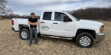 A man stands with his arms crossed next to a white Chevrolet Silverado 2500 HD pickup truck parked in a dry, grassy field. The man wears a black t-shirt, blue jeans, and a baseball cap, smiling at the camera. The front door of the truck features a custom logo that reads "Jacks Lawn Service" along with a phone number. In the background, a line of bare trees sits under an overcast sky with textured white and gray clouds.