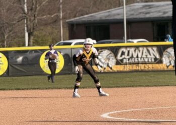 A youth softball player in a black, gold, and white uniform stands in a defensive ready position on a dirt infield. In the background, an outfield fence features a banner reading "Cowan Blackhawks Softball" alongside an American flag flying on a tall flagpole.
