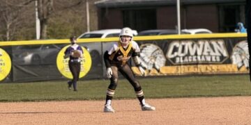 A youth softball player in a black, gold, and white uniform stands in a defensive ready position on a dirt infield. In the background, an outfield fence features a banner reading "Cowan Blackhawks Softball" alongside an American flag flying on a tall flagpole.