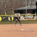 A youth softball player in a black, gold, and white uniform stands in a defensive ready position on a dirt infield. In the background, an outfield fence features a banner reading "Cowan Blackhawks Softball" alongside an American flag flying on a tall flagpole.