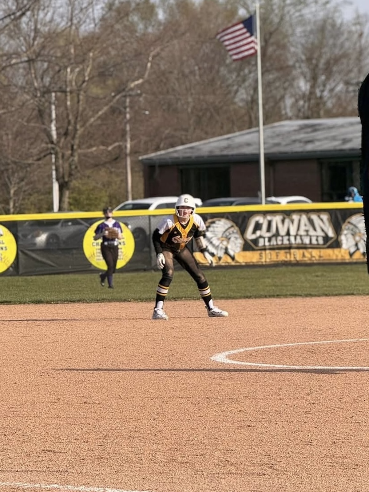 A youth softball player in a black, gold, and white uniform stands in a defensive ready position on a dirt infield. In the background, an outfield fence features a banner reading "Cowan Blackhawks Softball" alongside an American flag flying on a tall flagpole.
