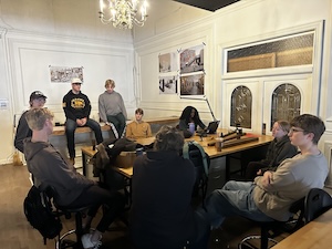A group of about nine people sit and stand around a long wooden table in a room with white walls and a chandelier. Some individuals are seated on chairs around the table, while others sit on a bench running along the back wall. Architectural design posters and renderings are pinned to the wall behind the table, and a glass-paneled double door is visible on the right.