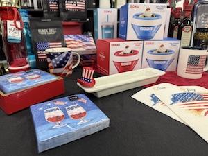 A retail merchandise display table covered with a black tablecloth, filled with patriotic American-themed home goods. In the foreground sit packs of paper napkins depicting layered red, white, and blue summer cocktails, alongside printed paper menus. A long, rectangular white ceramic serving dish sits in the center with a small Uncle Sam top hat decoration perched on the rim. In the background are American flag-patterned mugs, retail boxes for Zoku ice cream makers, and packages of Black Rifle Coffee.