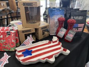 A close-up view of an indoor retail display table covered with a black tablecloth, featuring American-themed products. In the foreground sits a ceramic serving platter shaped like the map of the United States, painted with red and white stripes and a single blue star. Stacked behind it are brown cardboard retail boxes for "BenShot" glassware, a package of floral red napkins, two red "Fletchers' Mill" wood pepper mills, and several bags of "Black Rifle Coffee Company" coffee beans.