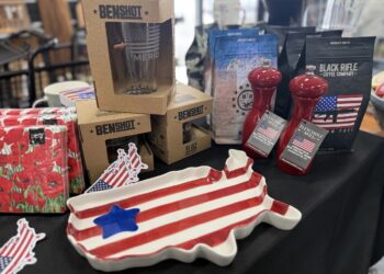 A close-up view of an indoor retail display table covered with a black tablecloth, featuring American-themed products. In the foreground sits a ceramic serving platter shaped like the map of the United States, painted with red and white stripes and a single blue star. Stacked behind it are brown cardboard retail boxes for "BenShot" glassware, a package of floral red napkins, two red "Fletchers' Mill" wood pepper mills, and several bags of "Black Rifle Coffee Company" coffee beans.