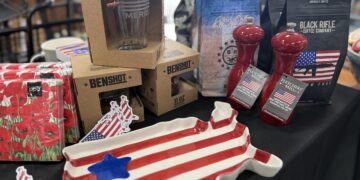 A close-up view of an indoor retail display table covered with a black tablecloth, featuring American-themed products. In the foreground sits a ceramic serving platter shaped like the map of the United States, painted with red and white stripes and a single blue star. Stacked behind it are brown cardboard retail boxes for "BenShot" glassware, a package of floral red napkins, two red "Fletchers' Mill" wood pepper mills, and several bags of "Black Rifle Coffee Company" coffee beans.