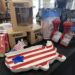 A close-up view of an indoor retail display table covered with a black tablecloth, featuring American-themed products. In the foreground sits a ceramic serving platter shaped like the map of the United States, painted with red and white stripes and a single blue star. Stacked behind it are brown cardboard retail boxes for "BenShot" glassware, a package of floral red napkins, two red "Fletchers' Mill" wood pepper mills, and several bags of "Black Rifle Coffee Company" coffee beans.