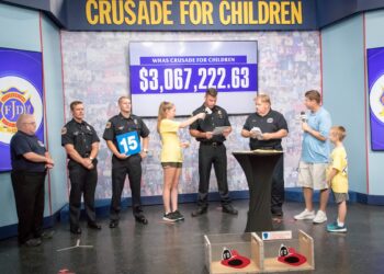 Firefighters from the Jeffersontown Fire Department present a donation on a television studio stage during the WHAS Crusade for Children telethon. A digital screen in the background displays a fundraising total of over three million dollars, while a presenter interviews a firefighter at a podium.
