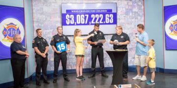 Firefighters from the Jeffersontown Fire Department present a donation on a television studio stage during the WHAS Crusade for Children telethon. A digital screen in the background displays a fundraising total of over three million dollars, while a presenter interviews a firefighter at a podium.
