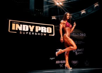 A female fitness competitor poses on a dark stage at the IFBB/NPC Indy Pro Supershow. She wears a sparkling red bikini and clear high-heeled shoes, showcasing her muscular physique while lifting one leg slightly. In the background, a large illuminated sign reads "IFBB/NPC Indy Pro Supershow," with various sponsor logos visible below it on a black backdrop.