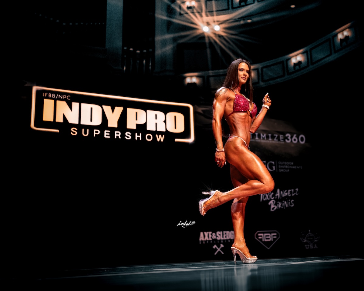 A female fitness competitor poses on a dark stage at the IFBB/NPC Indy Pro Supershow. She wears a sparkling red bikini and clear high-heeled shoes, showcasing her muscular physique while lifting one leg slightly. In the background, a large illuminated sign reads "IFBB/NPC Indy Pro Supershow," with various sponsor logos visible below it on a black backdrop.