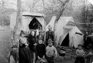 A monochrome historical photograph captures a group of seven individuals, including men, women, and children, posed in a wooded campsite. In the background stands a horse-drawn wagon with a light-colored canvas arched cover, next to a large canvas wedge tent. A metal kettle sits on a small stand over an outdoor cooking spot in the dirt. The individuals are dressed in historical, layered clothing, with some wearing hats and headscarves, surrounded by bare trees.