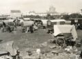 A monochrome historical photograph captures a large gathering of traditional wooden caravans and canvas-covered wagons parked across an open, grassy field. Several horses stand near the wagons, and a small dog rests on the ground in the foreground. Scattered around the campsite are household items, including chairs, buckets, and textiles. In the background, a small village stretches across the horizon, dominated by a large, medieval-style stone church tower under a pale sky. A small number "5" is printed in white in the bottom right corner.