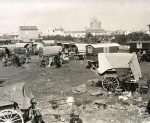 A monochrome historical photograph captures a large gathering of traditional wooden caravans and canvas-covered wagons parked across an open, grassy field. Several horses stand near the wagons, and a small dog rests on the ground in the foreground. Scattered around the campsite are household items, including chairs, buckets, and textiles. In the background, a small village stretches across the horizon, dominated by a large, medieval-style stone church tower under a pale sky. A small number "5" is printed in white in the bottom right corner.