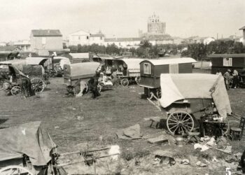 A monochrome historical photograph captures a large gathering of traditional wooden caravans and canvas-covered wagons parked across an open, grassy field. Several horses stand near the wagons, and a small dog rests on the ground in the foreground. Scattered around the campsite are household items, including chairs, buckets, and textiles. In the background, a small village stretches across the horizon, dominated by a large, medieval-style stone church tower under a pale sky. A small number "5" is printed in white in the bottom right corner.