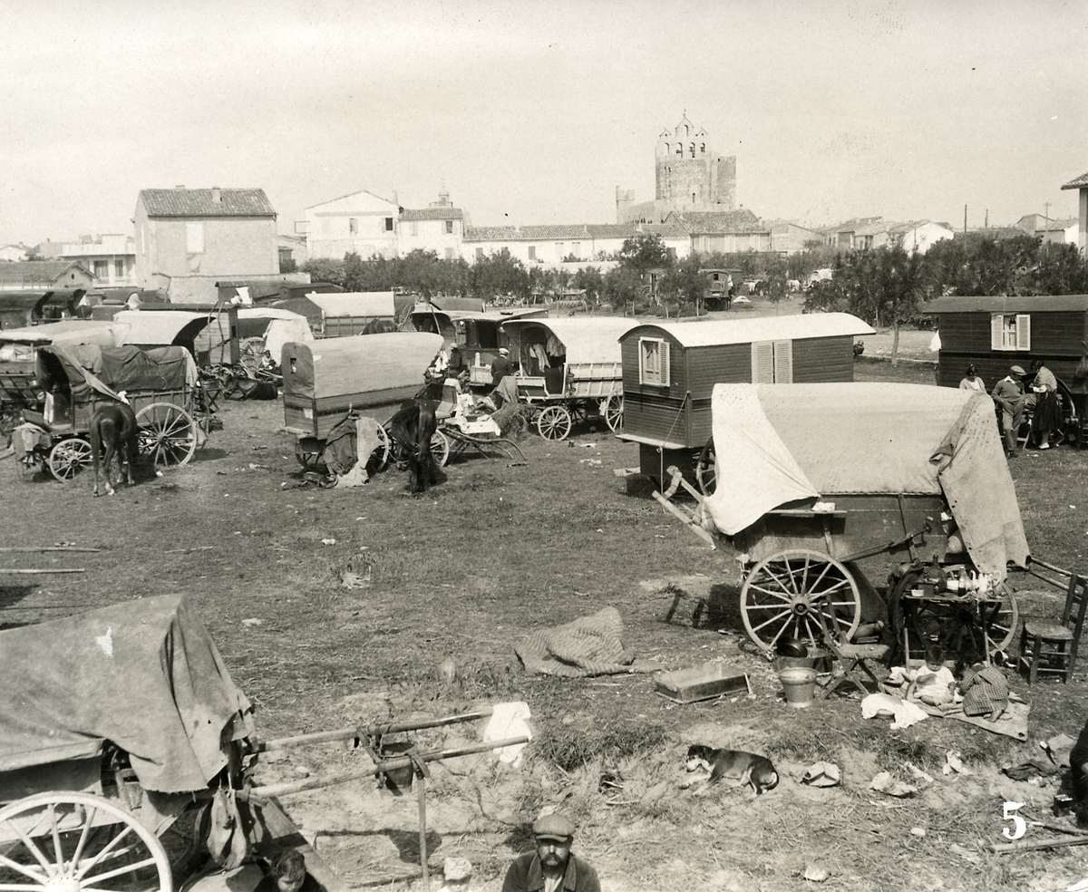A monochrome historical photograph captures a large gathering of traditional wooden caravans and canvas-covered wagons parked across an open, grassy field. Several horses stand near the wagons, and a small dog rests on the ground in the foreground. Scattered around the campsite are household items, including chairs, buckets, and textiles. In the background, a small village stretches across the horizon, dominated by a large, medieval-style stone church tower under a pale sky. A small number "5" is printed in white in the bottom right corner.