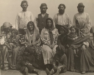 A vintage, sepia-toned studio photograph depicts an extended family group arranged in three rows against a plain backdrop. In the back row, four bearded men stand wearing traditional tunics, shirts, and one waistcoat. The middle row features seated women and children; two women wear traditional headscarves and patterned clothing, one holds an infant, and another man on the far right sits holding a young child. Two small children sit directly on the floor in the foreground.