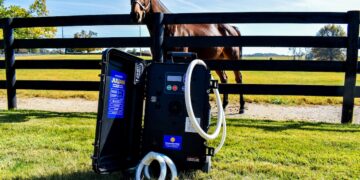 An eye-level shot of an equine therapeutic device, designed as a black portable hard case with white hoses, resting upright on a green grass lawn. The inside lid of the open case features a blue label with text reading "JULIAN." In the background, a brown horse wearing a halter stands behind a black three-rail wooden fence in a sunny paddock. The green pasture stretches toward a horizon lined with trees under a bright blue sky with light clouds.