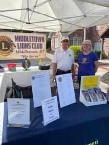 A man in a white polo shirt and a woman in a purple shirt stand behind promotional tables under a white canopy tent. A banner behind them reads "Middletown Lions Club, Middletown, KY, 'We Serve.'" Informational signs and brochures are arranged on the tables in the foreground.