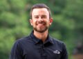 A head-and-shoulders portrait of a smiling young man with a neatly groomed beard and dark hair slicked back. He is wearing a black polo shirt featuring a white, stylized wing or mountain logo on the left chest. The background is a soft, blurred green, suggesting an outdoor setting like a park or wooded area.