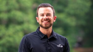 A head-and-shoulders portrait of a smiling young man with a neatly groomed beard and dark hair slicked back. He is wearing a black polo shirt featuring a white, stylized wing or mountain logo on the left chest. The background is a soft, blurred green, suggesting an outdoor setting like a park or wooded area.