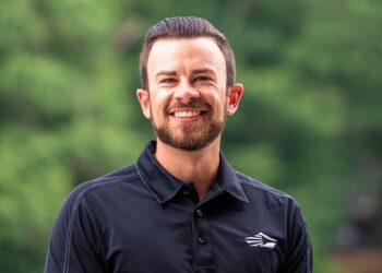 A head-and-shoulders portrait of a smiling young man with a neatly groomed beard and dark hair slicked back. He is wearing a black polo shirt featuring a white, stylized wing or mountain logo on the left chest. The background is a soft, blurred green, suggesting an outdoor setting like a park or wooded area.
