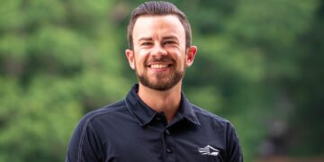 A head-and-shoulders portrait of a smiling young man with a neatly groomed beard and dark hair slicked back. He is wearing a black polo shirt featuring a white, stylized wing or mountain logo on the left chest. The background is a soft, blurred green, suggesting an outdoor setting like a park or wooded area.