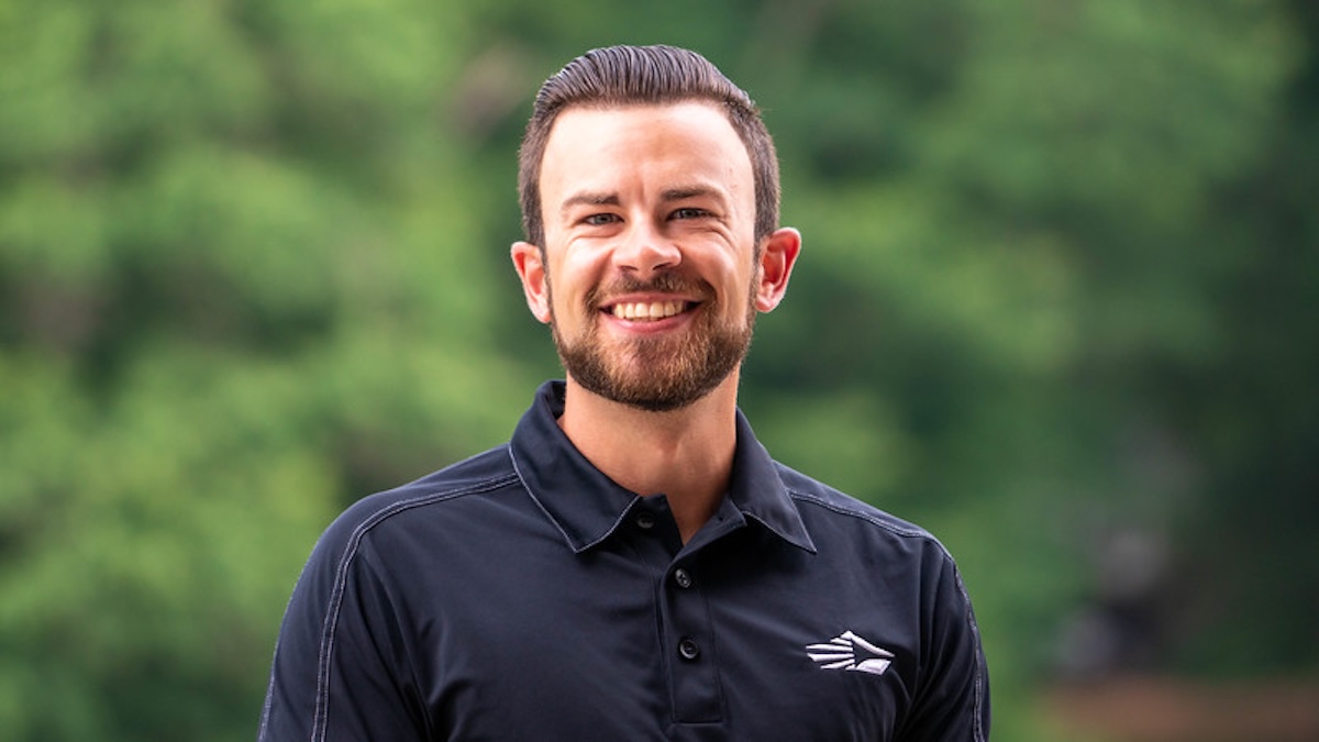 A head-and-shoulders portrait of a smiling young man with a neatly groomed beard and dark hair slicked back. He is wearing a black polo shirt featuring a white, stylized wing or mountain logo on the left chest. The background is a soft, blurred green, suggesting an outdoor setting like a park or wooded area.