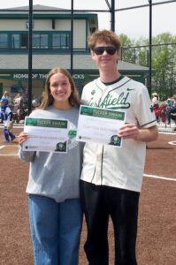 A young woman in a grey sweatshirt and a young man in a white and green Westfield baseball jersey stand together on a dirt baseball field. They are both smiling and holding "Tucker Swain Memorial Scholarship" certificates. A green building with "HOME OF THE SHAMROCKS" written on it is visible in the background.