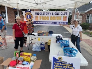 Three volunteers stand under a white canopy tent holding a large banner that reads "Middletown Lions Club Food Drive, Sunday, September 24, 1:00-5:00 PM." Folding tables display collection baskets, a prize wheel, and boxes filled with donated non-perishable food items.