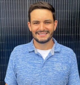 A portrait of a smiling man with a neat beard and short dark hair wearing a light blue heather polo shirt embroidered with a "Solar" company logo. He stands against a dark background composed of vertical lines resembling the surface of a solar panel.