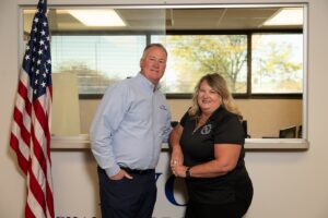 A medium shot of a man and a woman posing together in an office setting. The man, on the left, wears a light blue button-down shirt and dark pants, with one hand in his pocket. The woman, on the right, wears a black polo shirt and smiles warmly, leaning slightly toward him. They stand in front of a wide interior window that looks into another office space with trees visible through a far window. To the far left, an American flag on a wooden pole is partially visible. At the bottom of the frame, the tops of large blue letters on the wall are cut off, suggesting a company or organizational name.