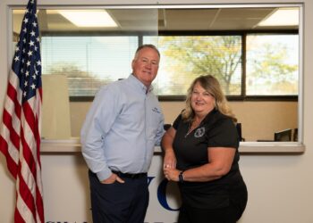 A medium shot of a man and a woman posing together in an office setting. The man, on the left, wears a light blue button-down shirt and dark pants, with one hand in his pocket. The woman, on the right, wears a black polo shirt and smiles warmly, leaning slightly toward him. They stand in front of a wide interior window that looks into another office space with trees visible through a far window. To the far left, an American flag on a wooden pole is partially visible. At the bottom of the frame, the tops of large blue letters on the wall are cut off, suggesting a company or organizational name.