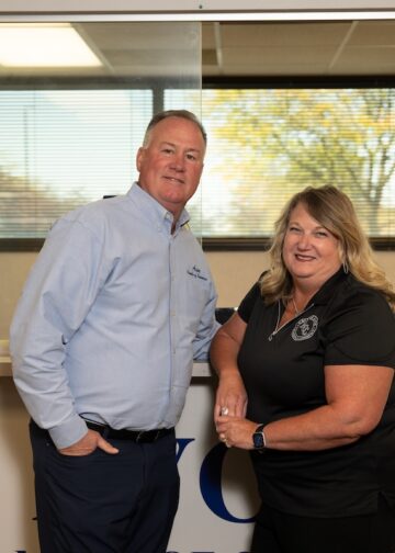 A medium shot of a man and a woman posing together in an office setting. The man, on the left, wears a light blue button-down shirt and dark pants, with one hand in his pocket. The woman, on the right, wears a black polo shirt and smiles warmly, leaning slightly toward him. They stand in front of a wide interior window that looks into another office space with trees visible through a far window. To the far left, an American flag on a wooden pole is partially visible. At the bottom of the frame, the tops of large blue letters on the wall are cut off, suggesting a company or organizational name.